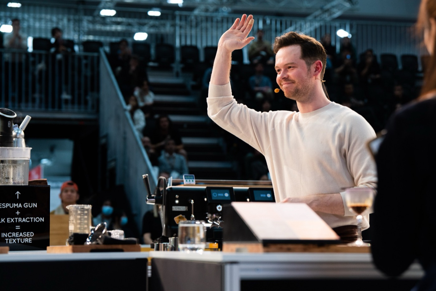 A barista waving to the crowd from a competition espresso station