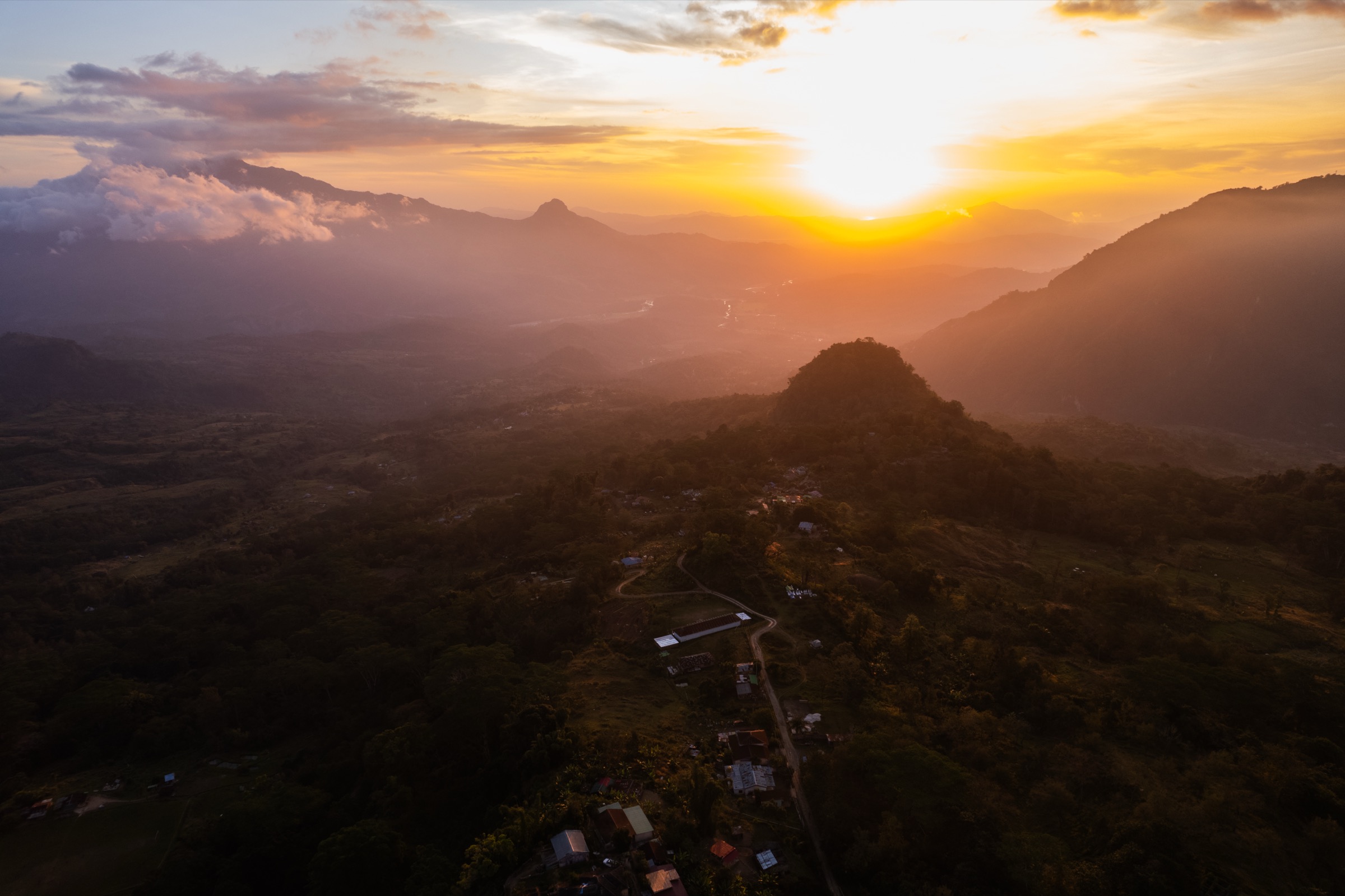 Aerial view at sunset over Timor-Leste: the sun going down behind mountain ridges, a village and a winding road on the forested hill below