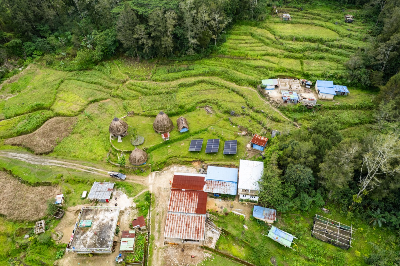 Aerial view of a hillside village with round thatched houses, three solar panels, tin roofs and green terraces