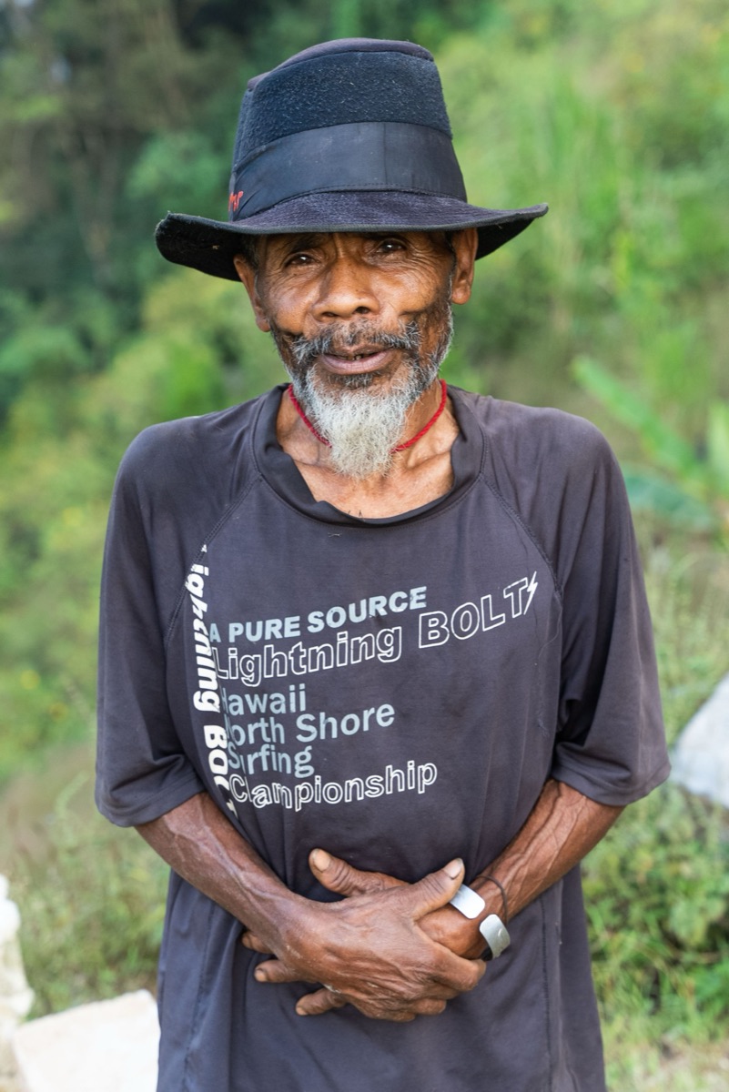 An older man with a grey beard, black hat and surf T-shirt, hands folded, in front of green hills