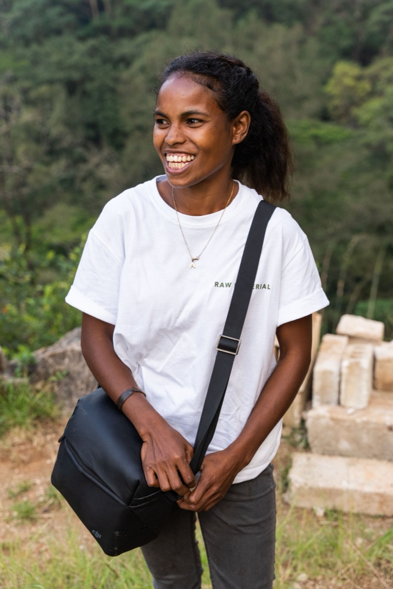 A young woman laughing, a camera bag over her shoulder, forest behind her