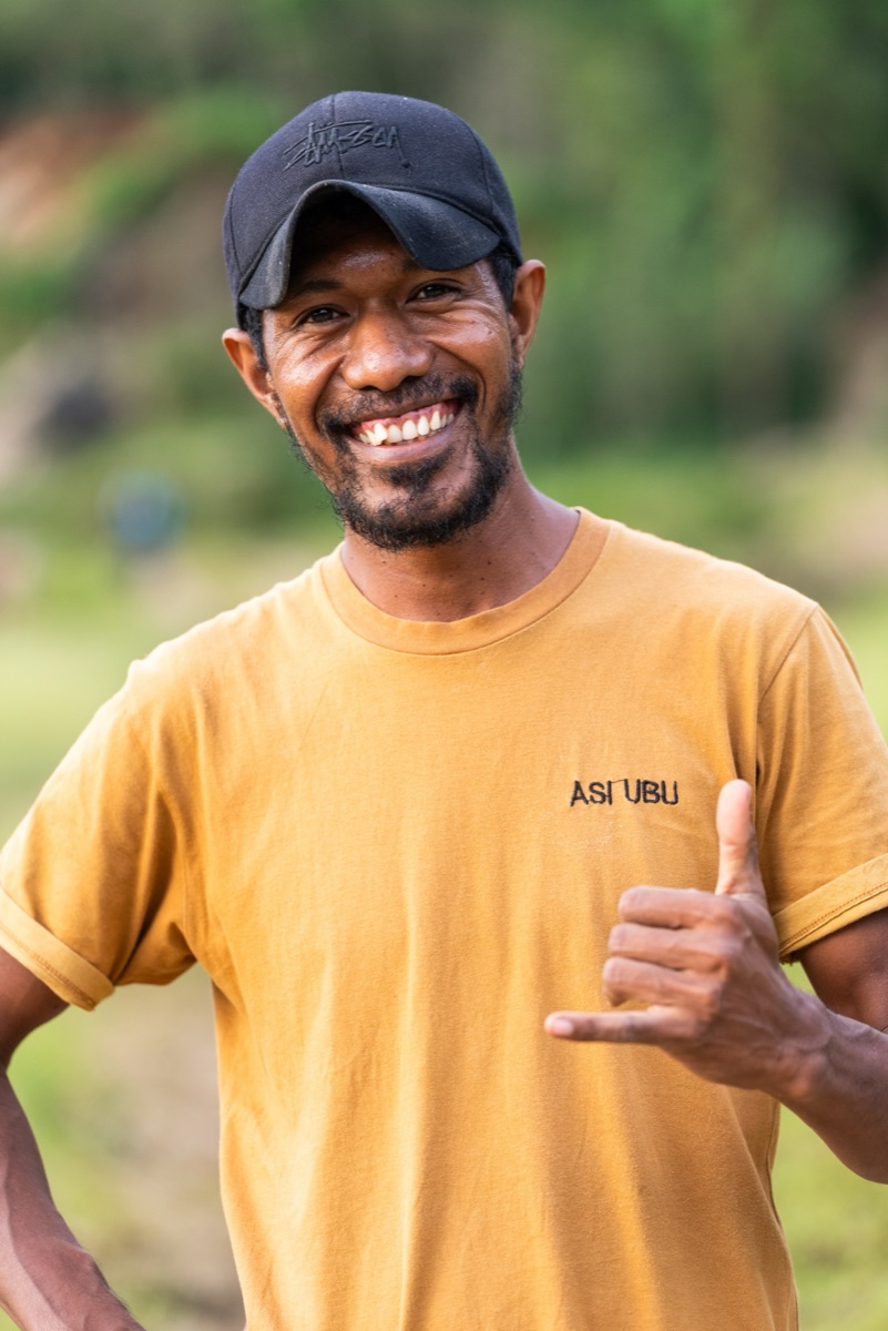 A young man in a mustard T-shirt and black cap grinning and throwing a shaka
